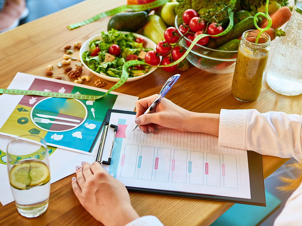 woman dietitian in medical uniform with tape measure working on a diet plan sitting with different healthy food ingredients in the green office on background. weight loss and right nutrition concept
